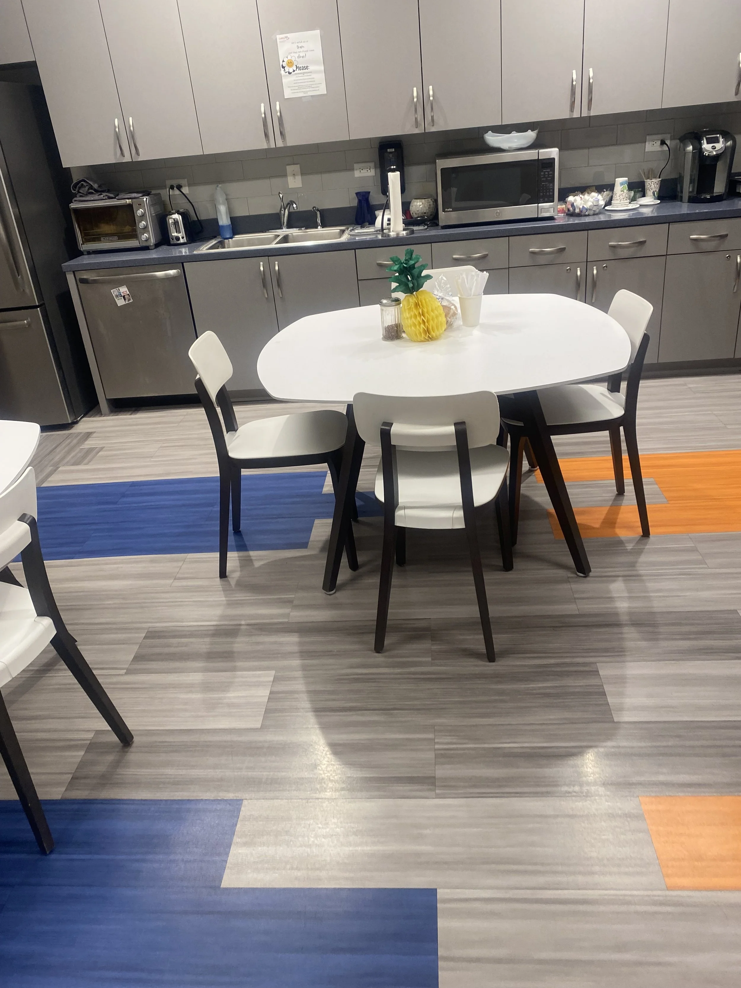 Kitchen with a white oval table surrounded by six chairs, a pineapple-shaped decor, and a countertop with appliances including a microwave, toaster, and coffee machine.