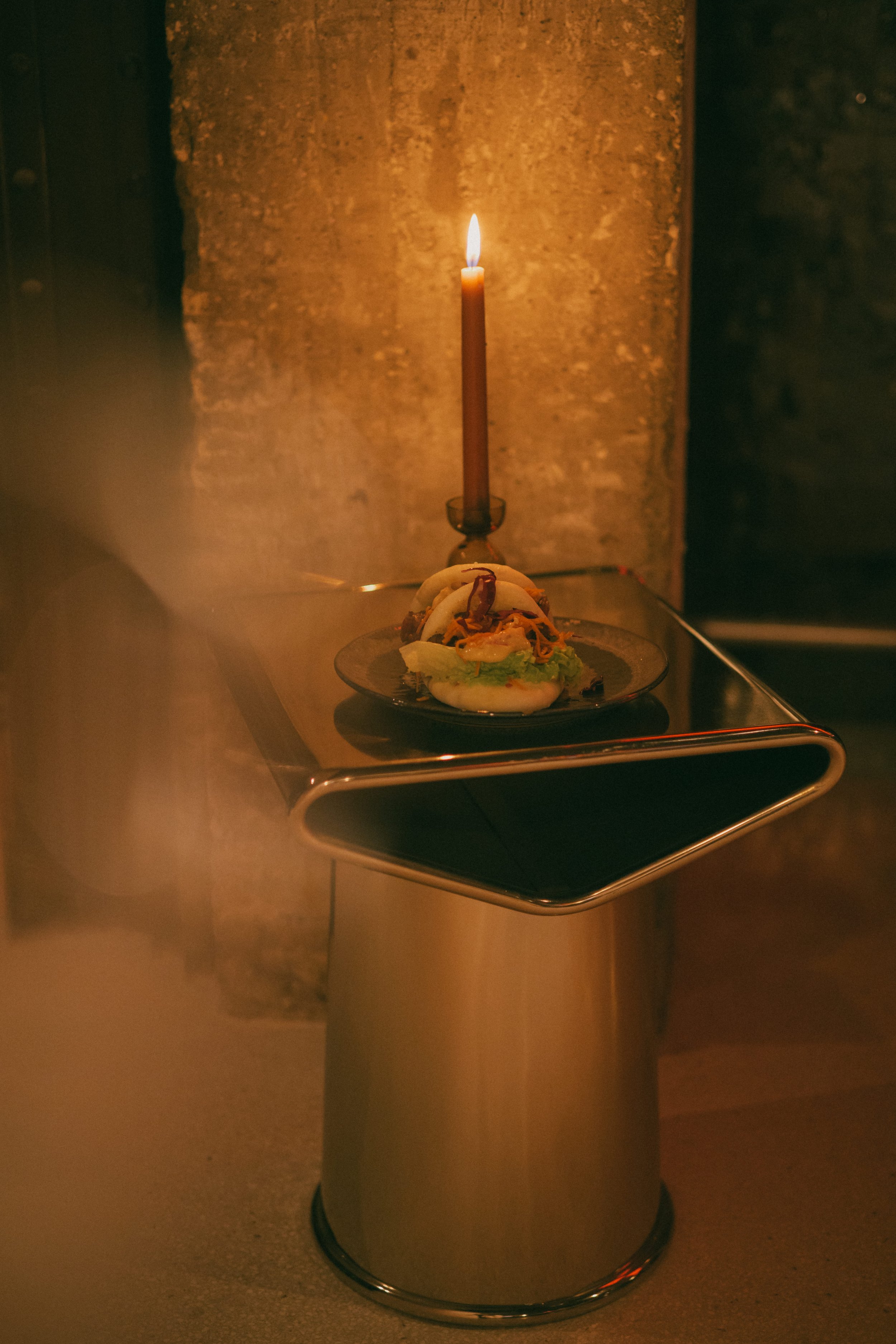 Une petite table en métal avec une assiette de nourriture, une bougie allumée sur un fond de mur en pierre.