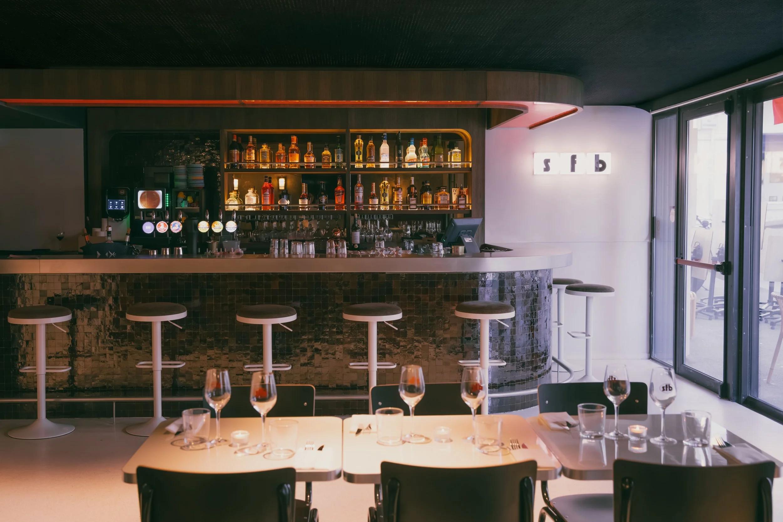 Bar intérieur avec comptoir en pierre, étagères de bouteilles d'alcool, tables avec verres de vin et bougie, grande fenêtre en vidro.