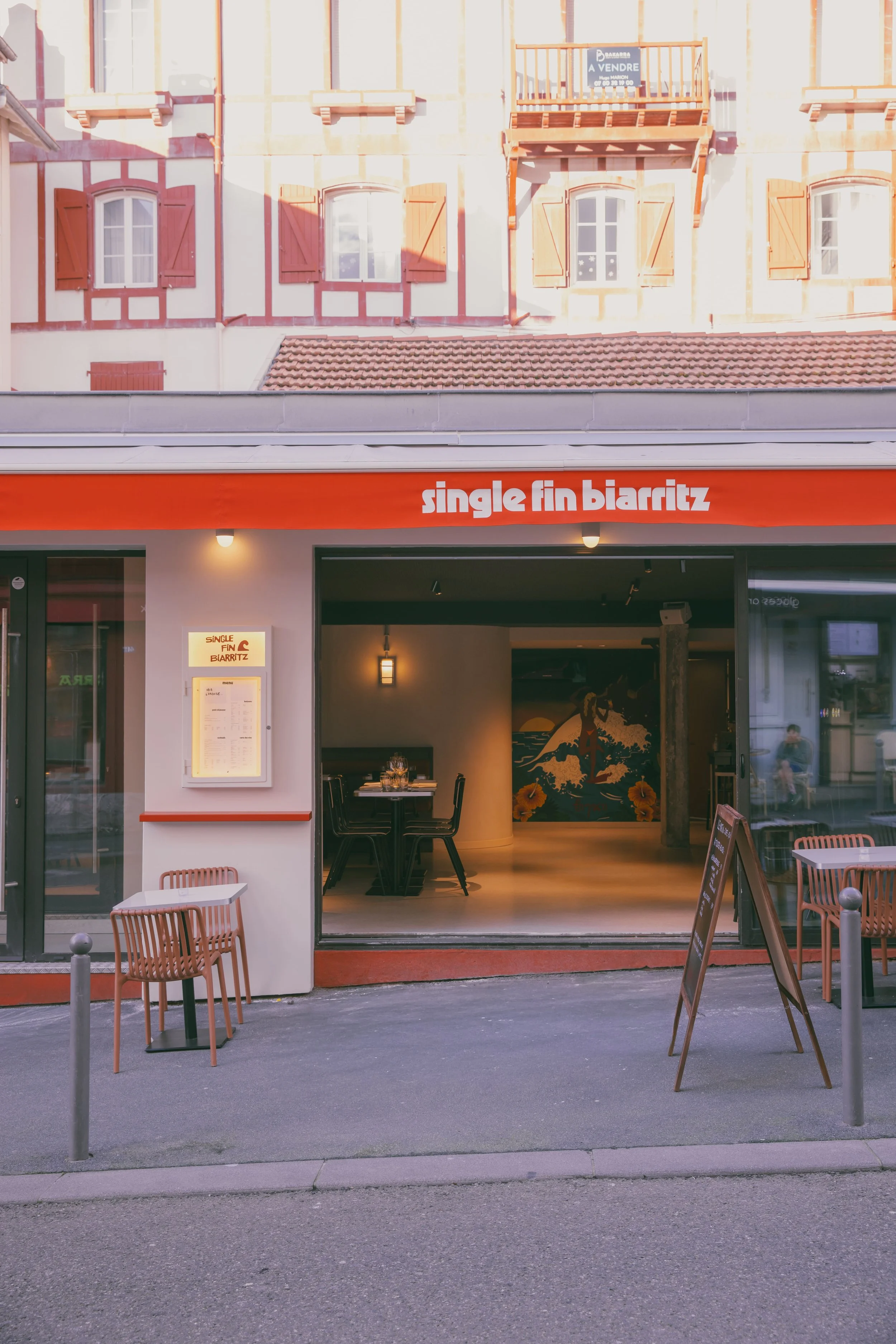 Façade d'un restaurant nommé 'single fin biarritz' avec une entrée ouverte, quelques tables et chaises à l'extérieur, tableau d'affichage à gauche, et un panneau de menu sur pied à droite.