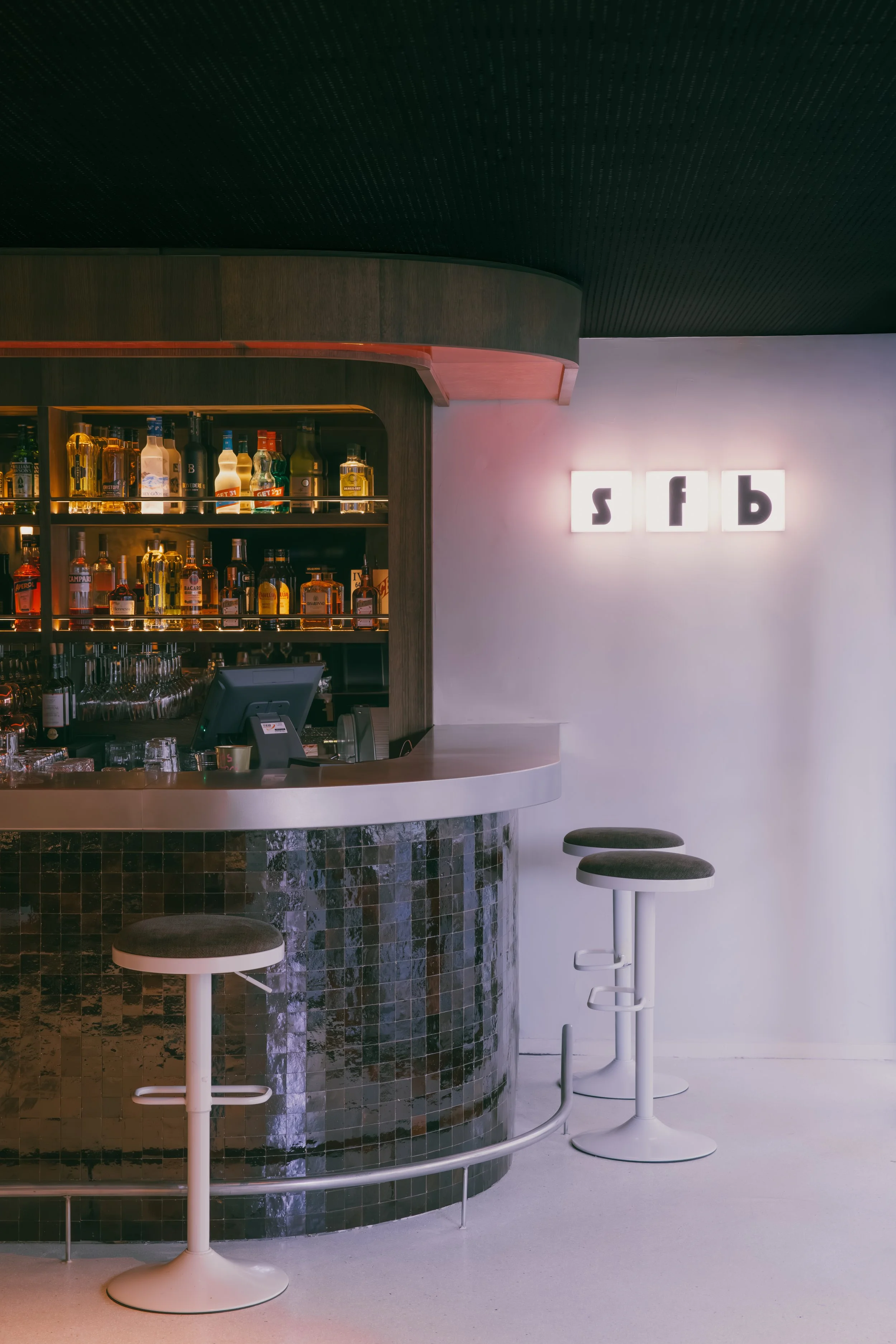 Bar avec comptoir en mosaïque sombre, étagères avec bouteilles de liquor, signal lumineux avec lettres ‘sfb’ sur le mur blanc