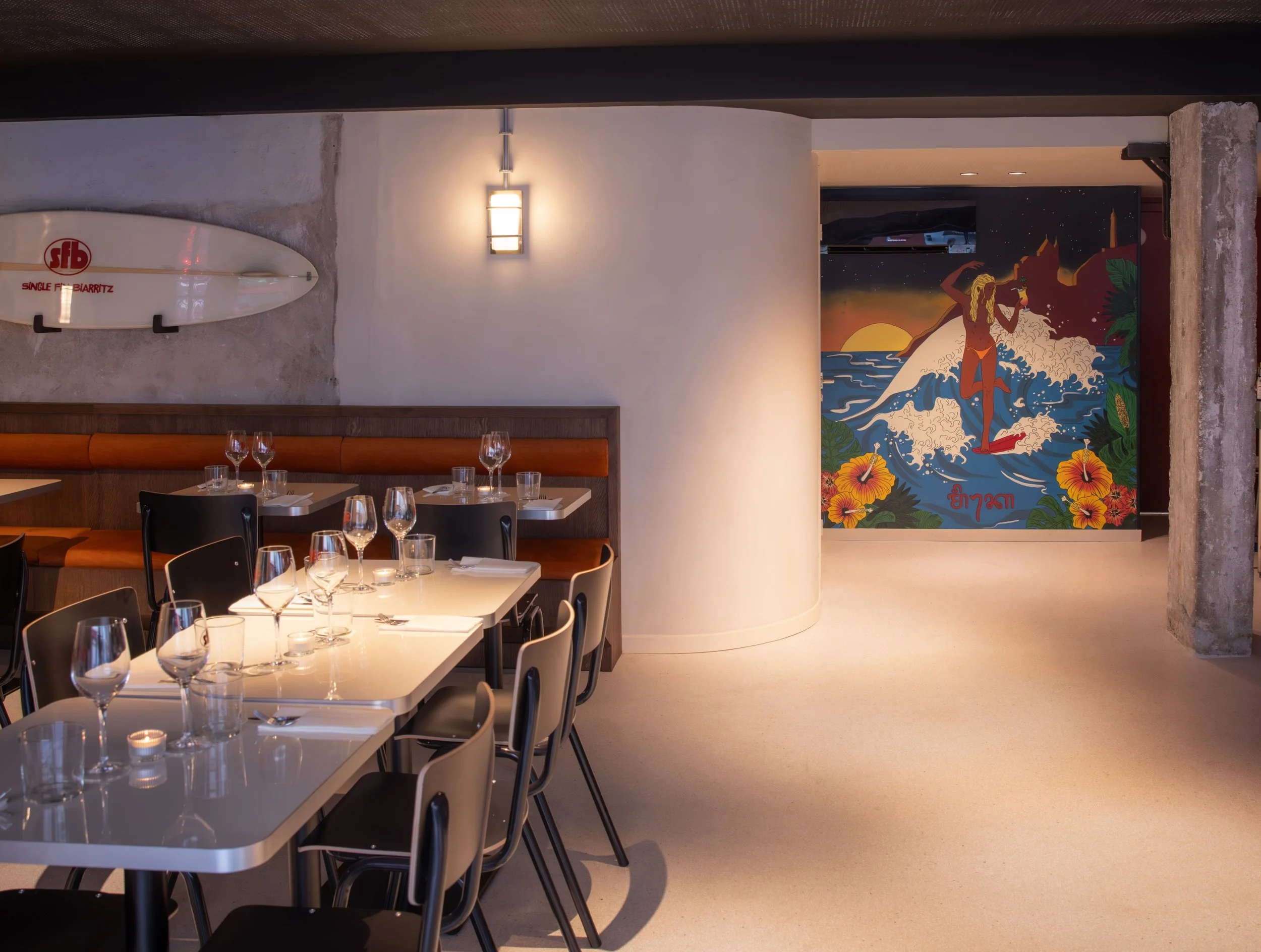 Salle à manger de restaurant avec tables, verres et assiettes, mur avec une planche de surf et une peinture murale colorée représentant une femme sur une vague, plage, soleil et fleurs.
