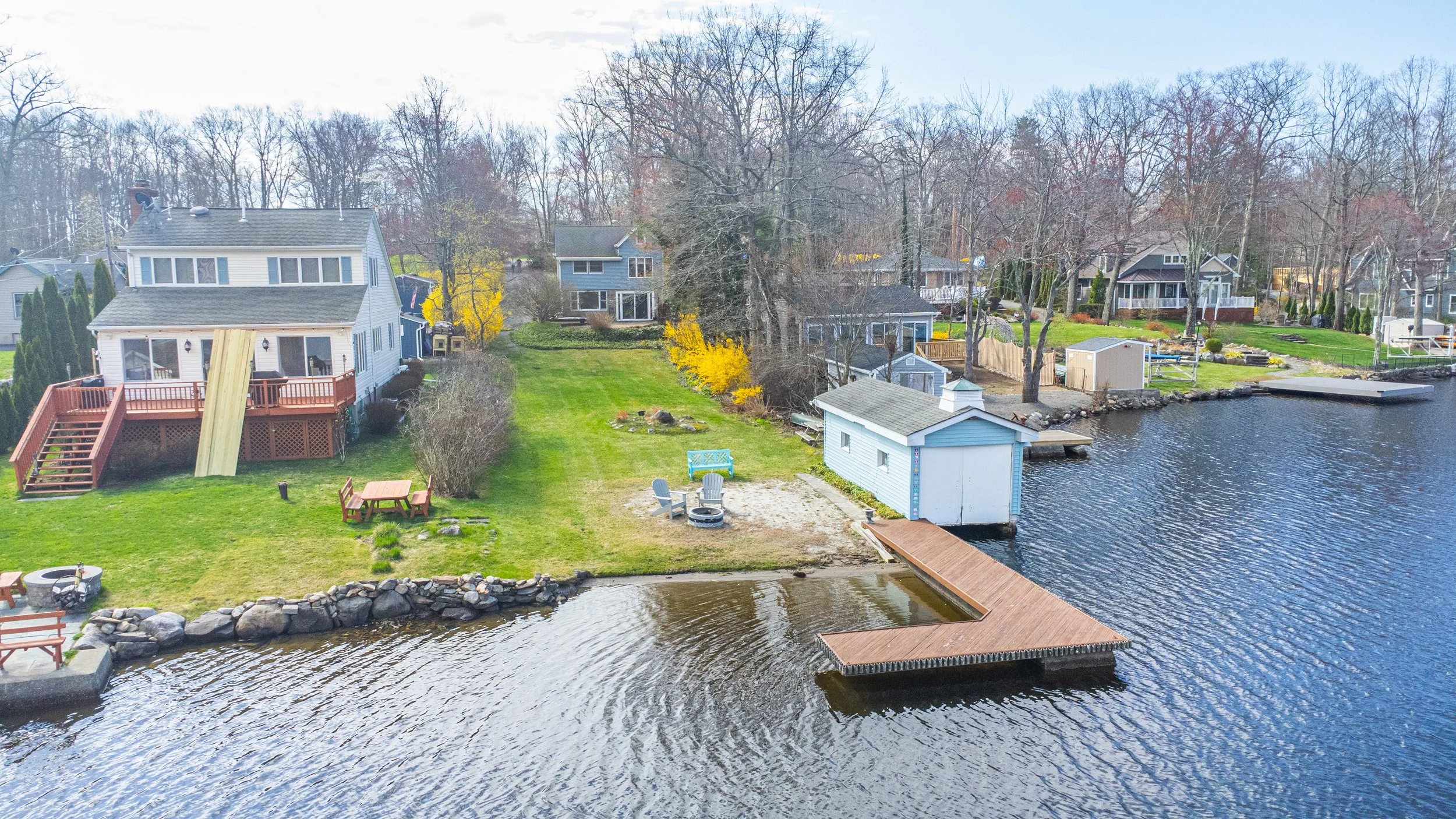Lakeside residential backyard with docks, trees, and houses, some with decks and outdoor furniture, under a cloudy sky.
