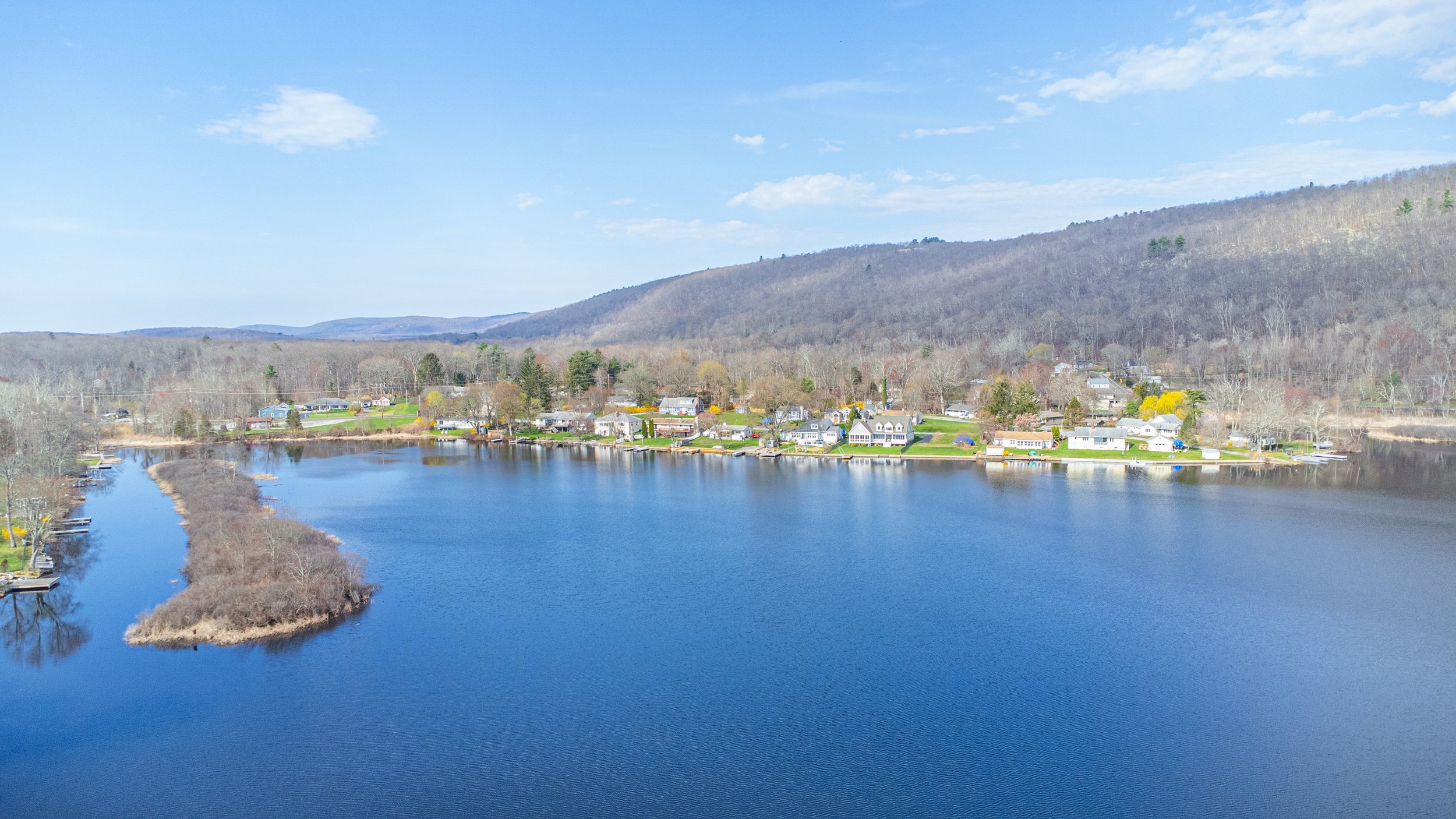 A scenic view of a small town along a river with houses, trees, and hills in the background under a partly cloudy sky.
