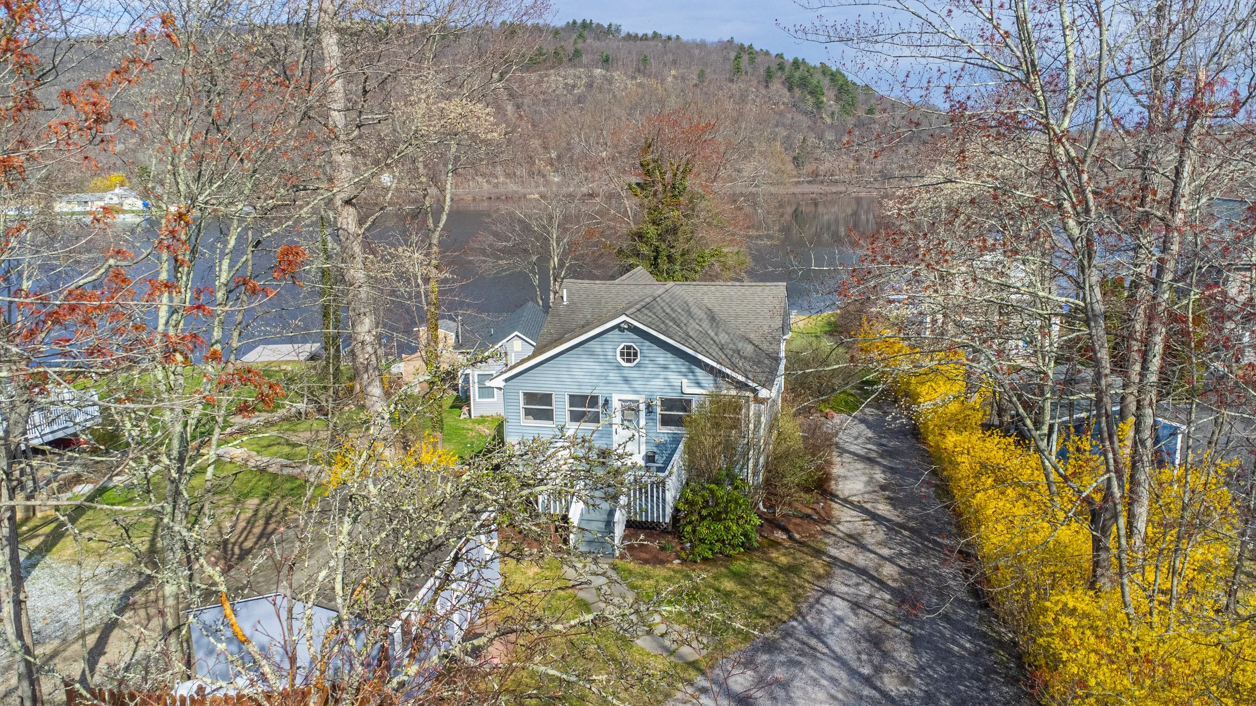 Bird's eye view of a house near a river, surrounded by trees with some colored leaves and bare branches, in a mountainous landscape on a clear day.
