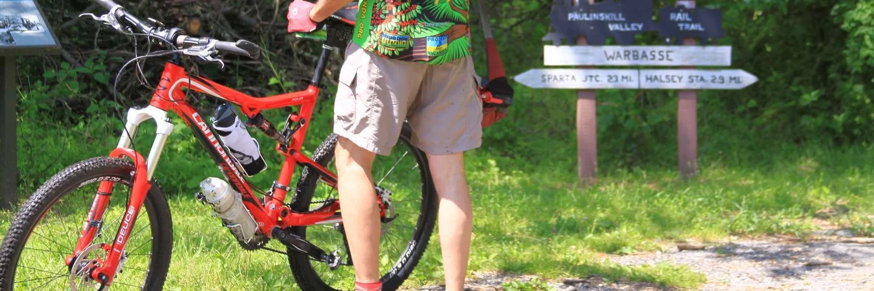 Person standing next to a red mountain bike on a grassy trail, holding a backpack, with trail signs in the background indicating directions to Warbasse and other locations.