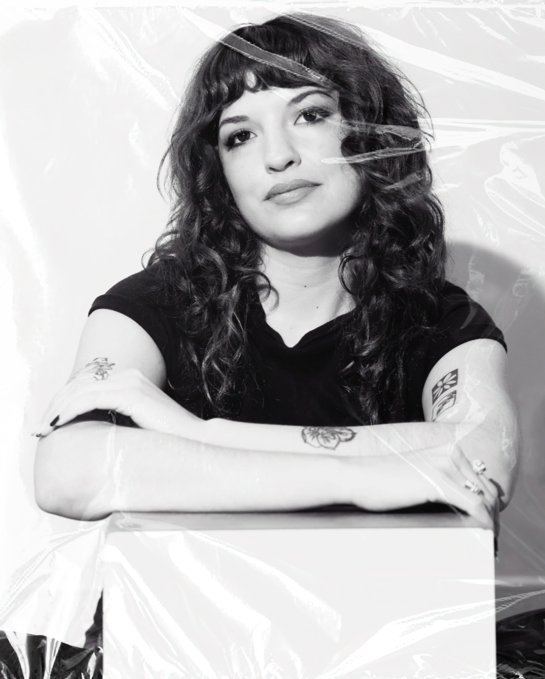 Haley Dani, a woman with curly hair, wearing a black shirt, sitting with arms crossed on a white surface, looking at the camera in a black-and-white photo.