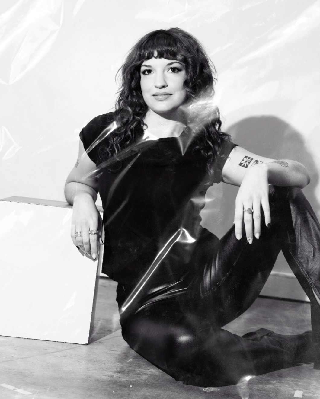 Haley Dani sitting on the floor with one arm resting on her knee, wearing dark clothing, with tattoos visible on her left arm, in front of a plain background.
