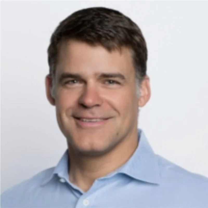 Headshot of a man with brown hair wearing a light blue collared shirt, smiling against a plain white background.