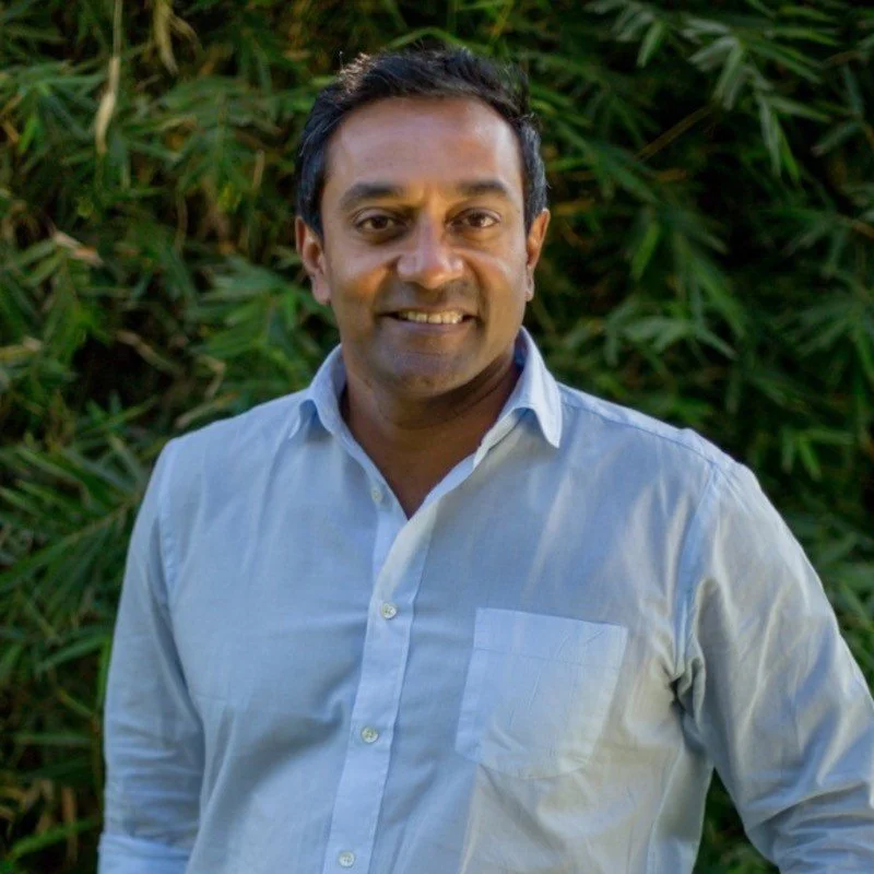 A man with dark hair and brown skin wearing a light blue button-up shirt, standing outdoors in front of green foliage, smiling at the camera.