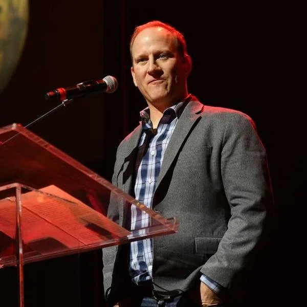 A man in a gray blazer and plaid shirt standing in front of a microphone on a stage, with a transparent podium nearby.