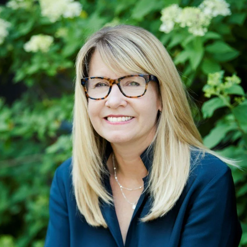 A smiling woman with blonde hair, glasses, and a black shirt, sitting outdoors with green foliage and white flowers in the background.