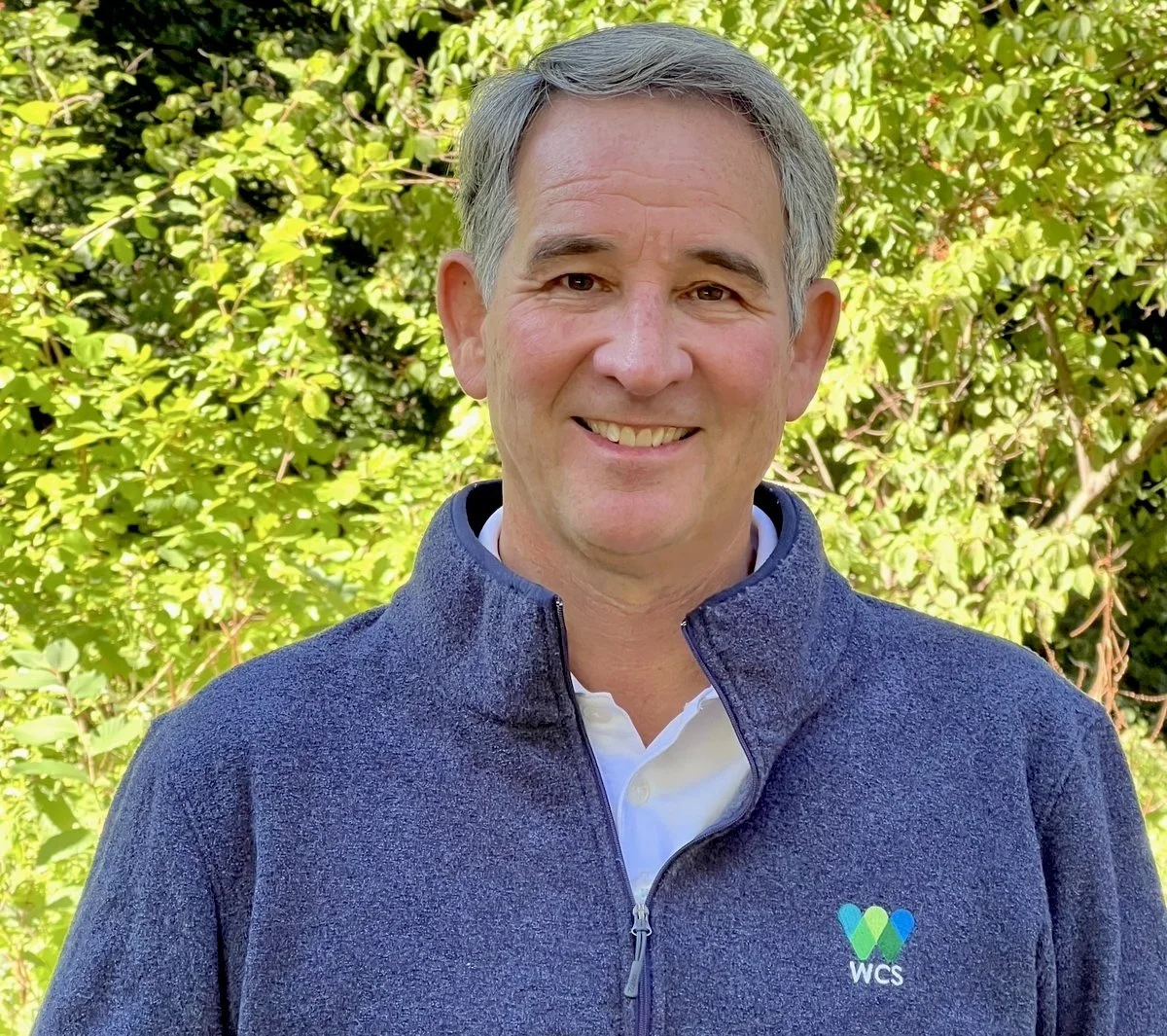 A smiling man with gray hair stands outdoors in front of green foliage, wearing a dark blue fleece jacket with a WCS logo.