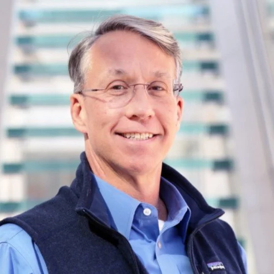 A smiling middle-aged man with glasses, wearing a blue button-up shirt and a dark vest, standing indoors with a blurred cityscape background visible through windows.