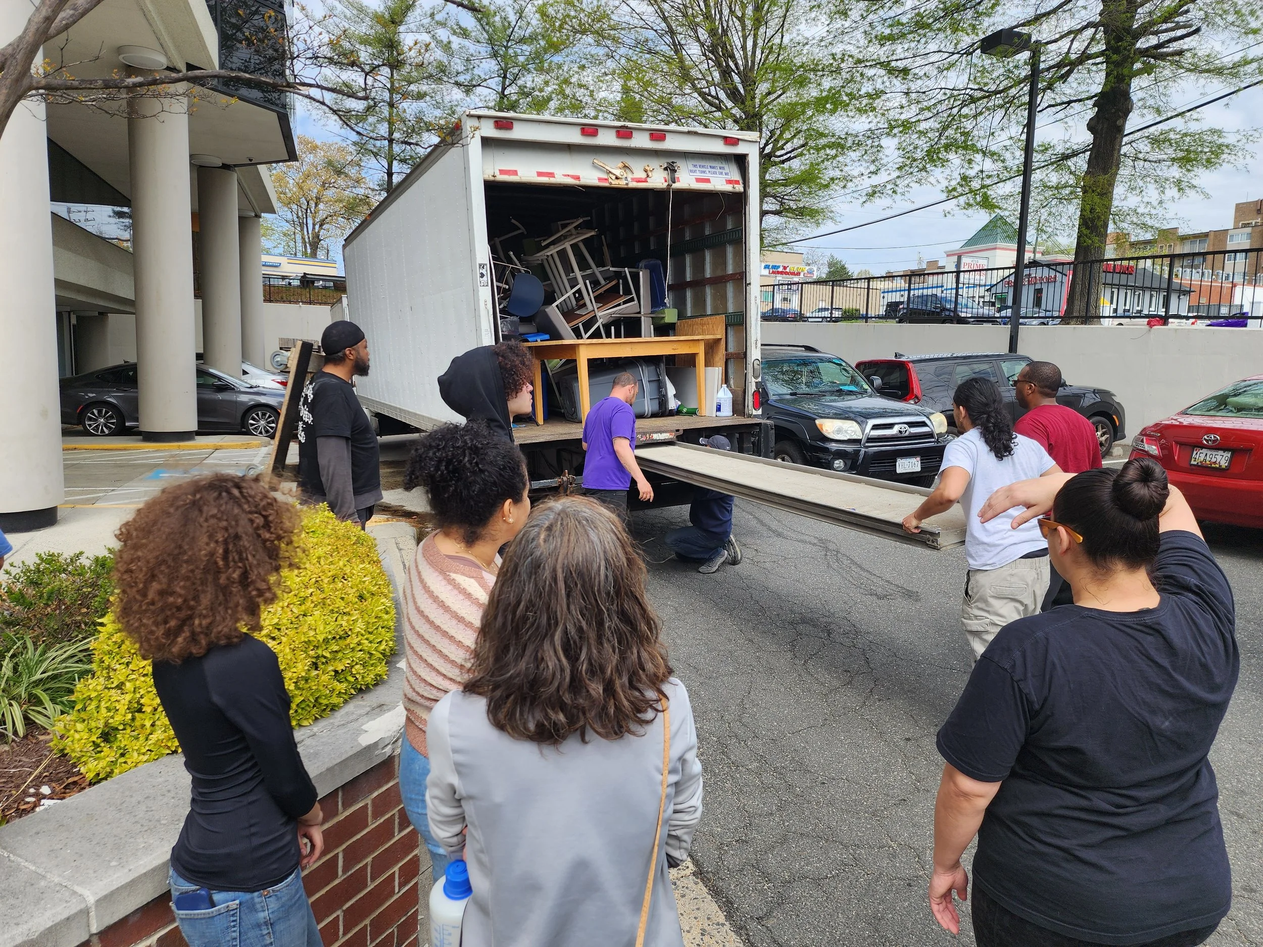 Group of people watching a moving truck unload furniture in a parking lot.
