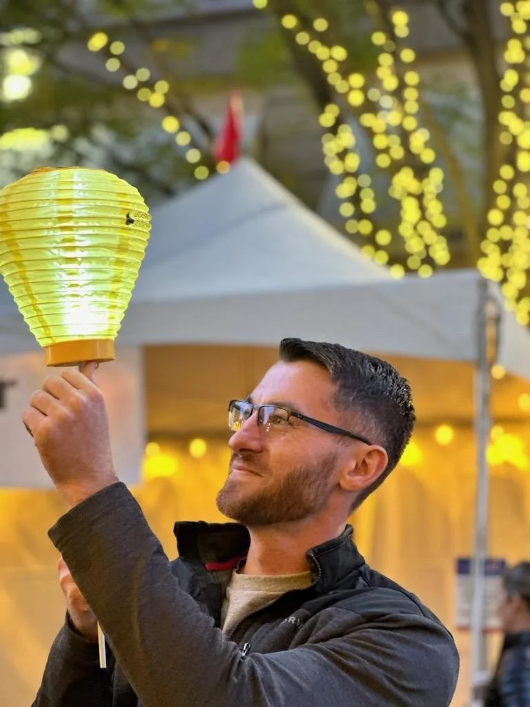 A man with glasses holding a yellow paper lantern at an outdoor event decorated with string lights.