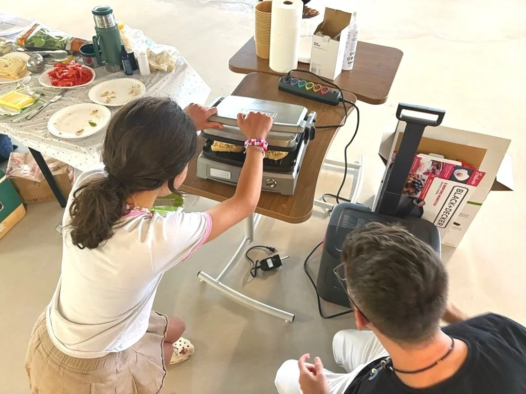 A girl using a sandwich press at a table, with a boy sitting nearby. The table has various food items, cups, and paper towels on it.