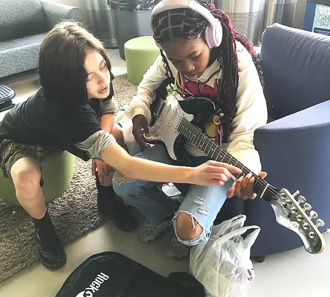 Two young girls sitting on the floor of a room, one with headphones playing an electric guitar while the other touches the guitar's strings, surrounded by furniture and a backpack.