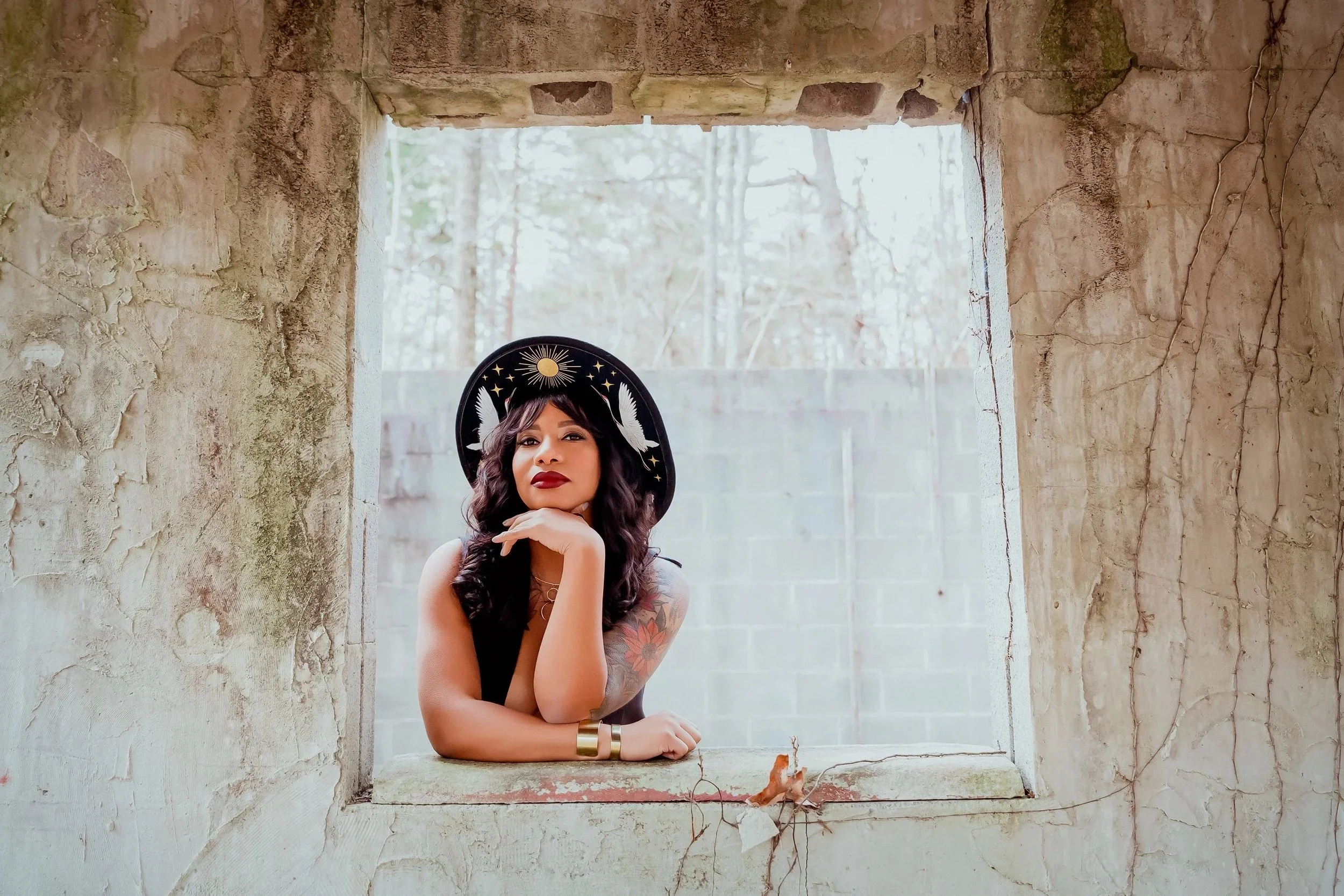 Jay poses in a concrete window frame with a hand under her chin. She is wearing a black hat with white cranes and gold details painted on the underside of the brim.