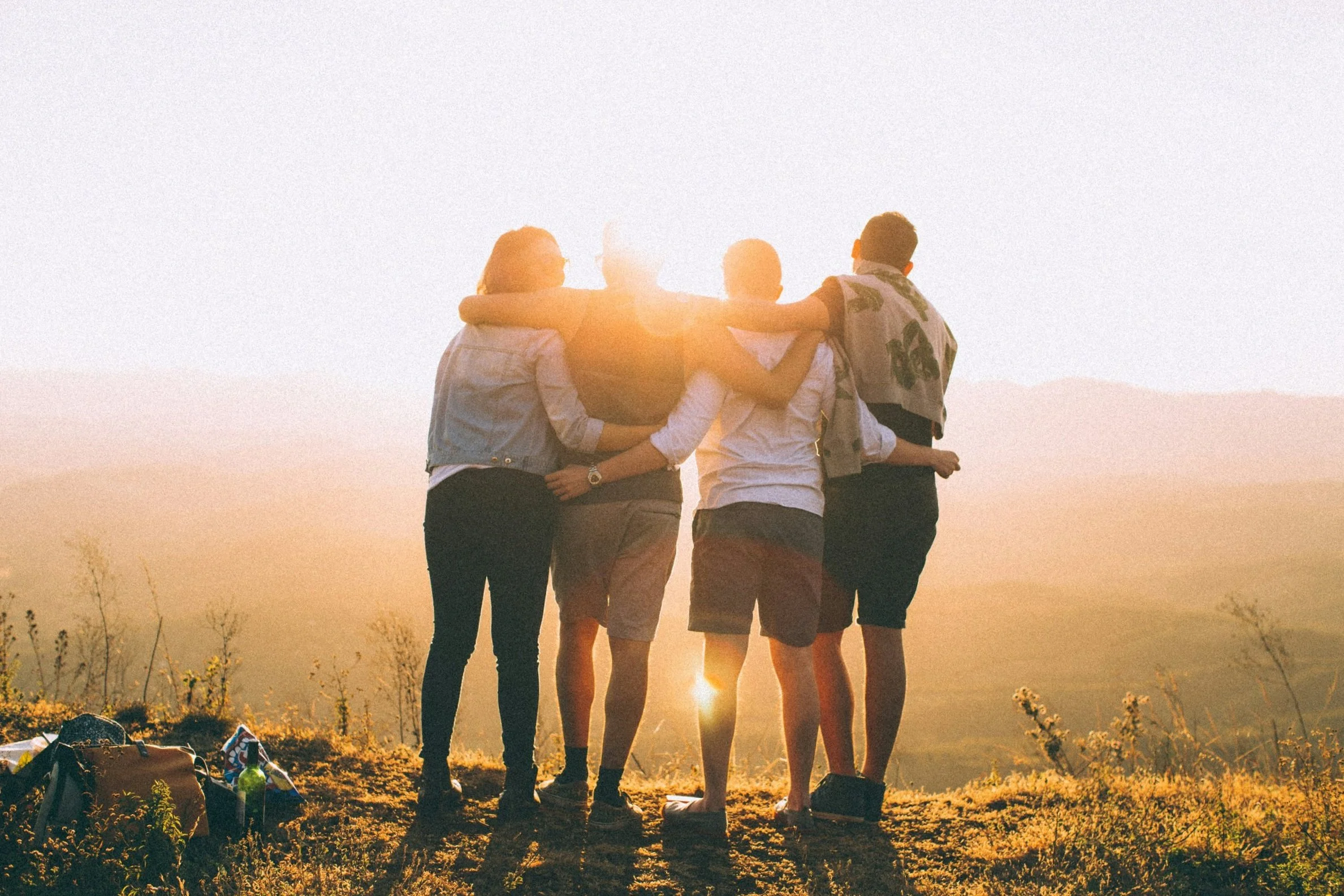 Groupe de cinq jeunes amis en tee-shirts et vêtements décontractés, marchant côte à côte avec bras autour des épaules, regardant vers un coucher de soleil sur une colline herbeuse pendant une belle journée.