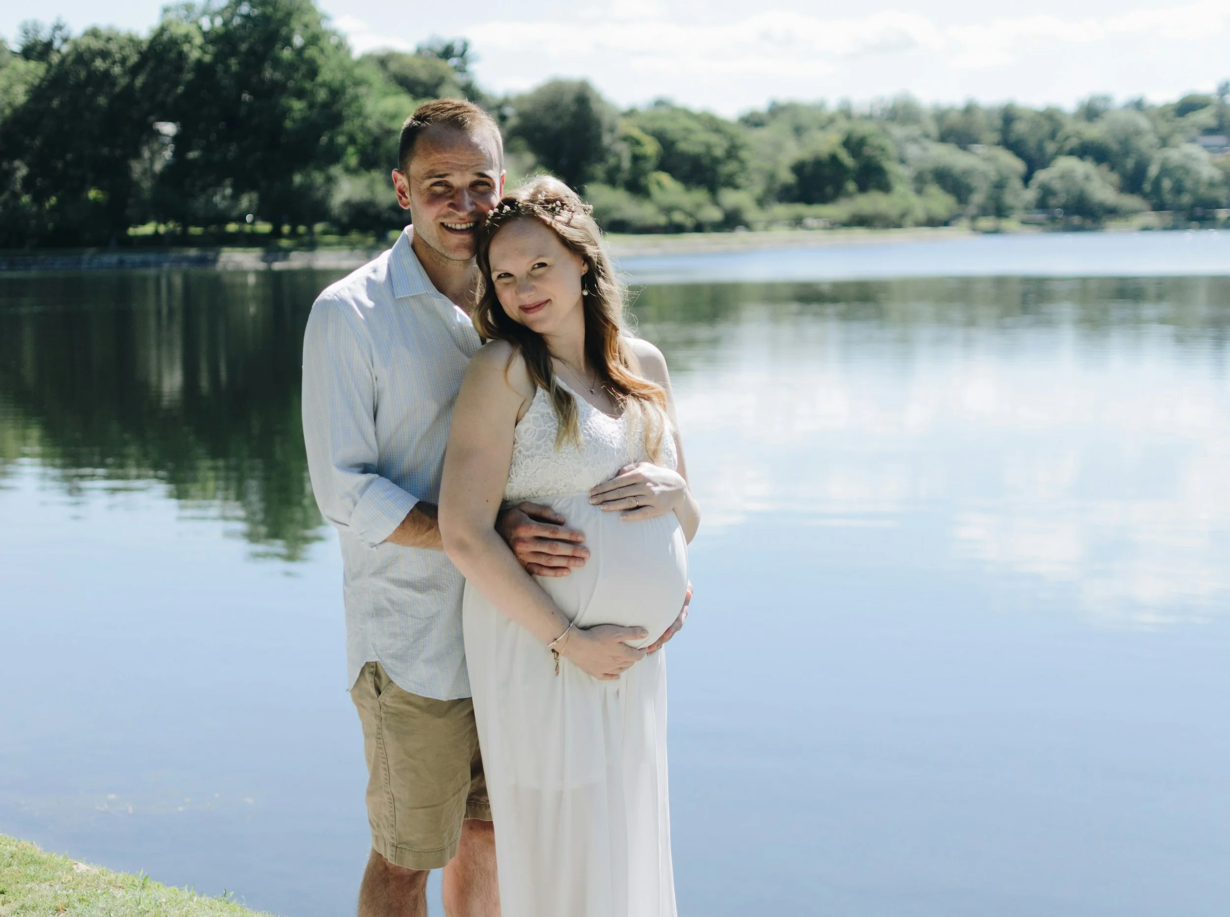 Un couple debout près d'un lac, la femme enceinte, tous deux souriants, entourés d'arbres et d'un ciel en partie nuageux.