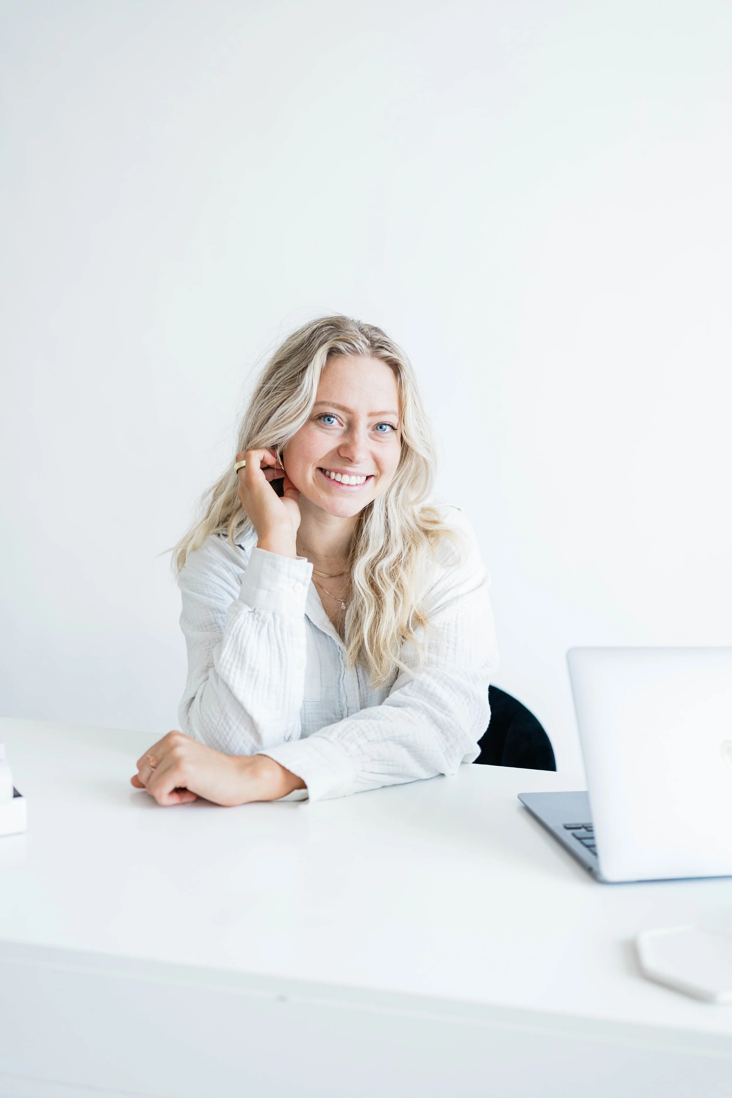 A young woman with blonde hair and blue eyes smiling, sitting at a white table with a silver laptop and a smartphone nearby, in a bright, minimalistic office or room with white walls.