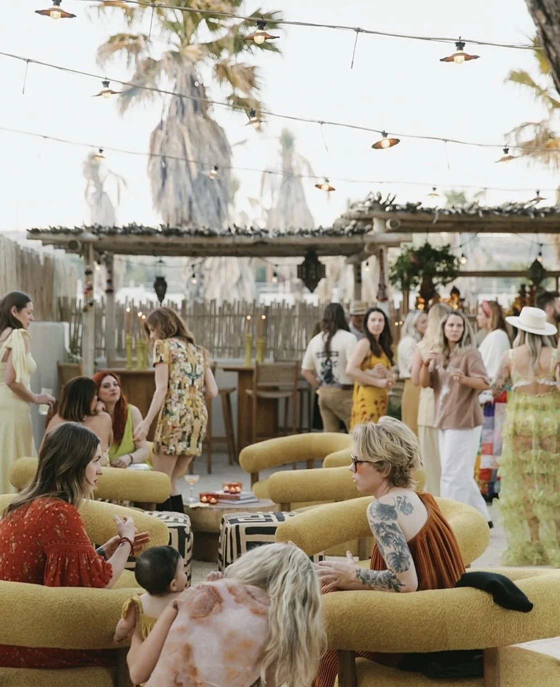 People gathered at an outdoor party with tropical decorations, string lights, and palm trees in the background, sitting on yellow couches and chatting.