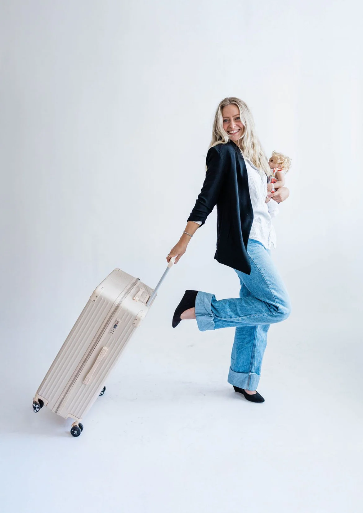 A smiling woman with blonde hair wearing jeans, a white shirt, a black jacket, and black heels, holding a doll in her left arm and pulling a beige suitcase with her right hand while standing on one leg against a plain white backdrop.