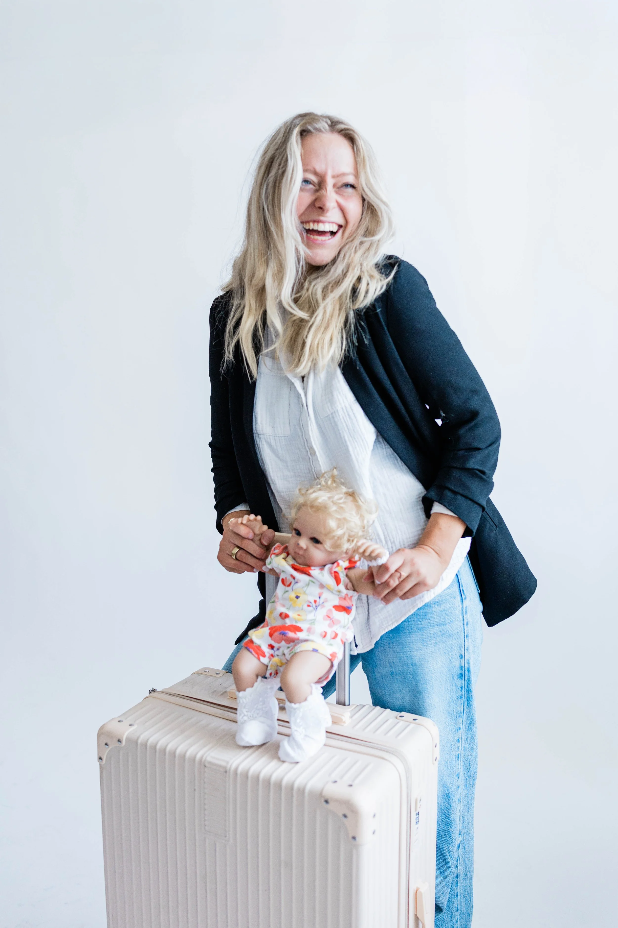 A smiling woman with blonde hair wearing a black blazer and light jeans holding a young girl with blonde curly hair, sitting on a cream-colored suitcase. The girl is dressed in a colorful outfit with white shoes.