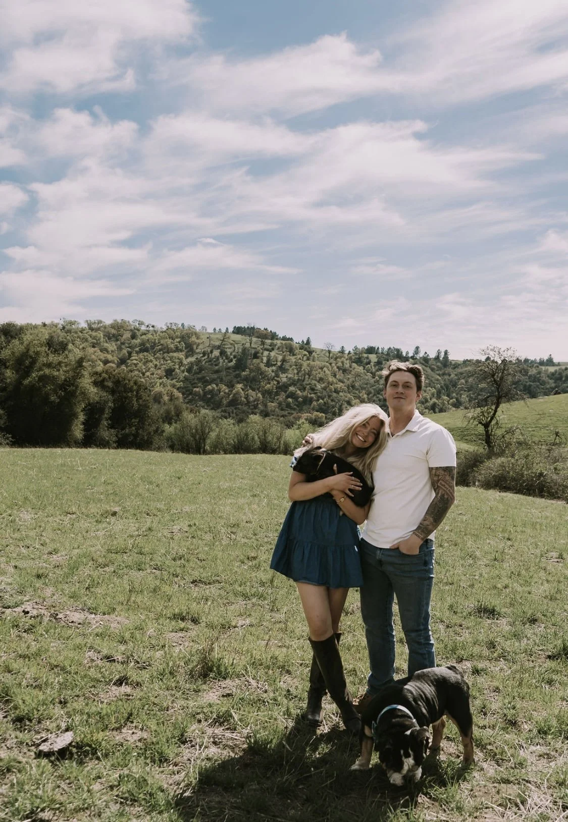 A happy couple standing outdoors in a grassy field with a small black and white dog, with rolling hills and a partly cloudy sky in the background.