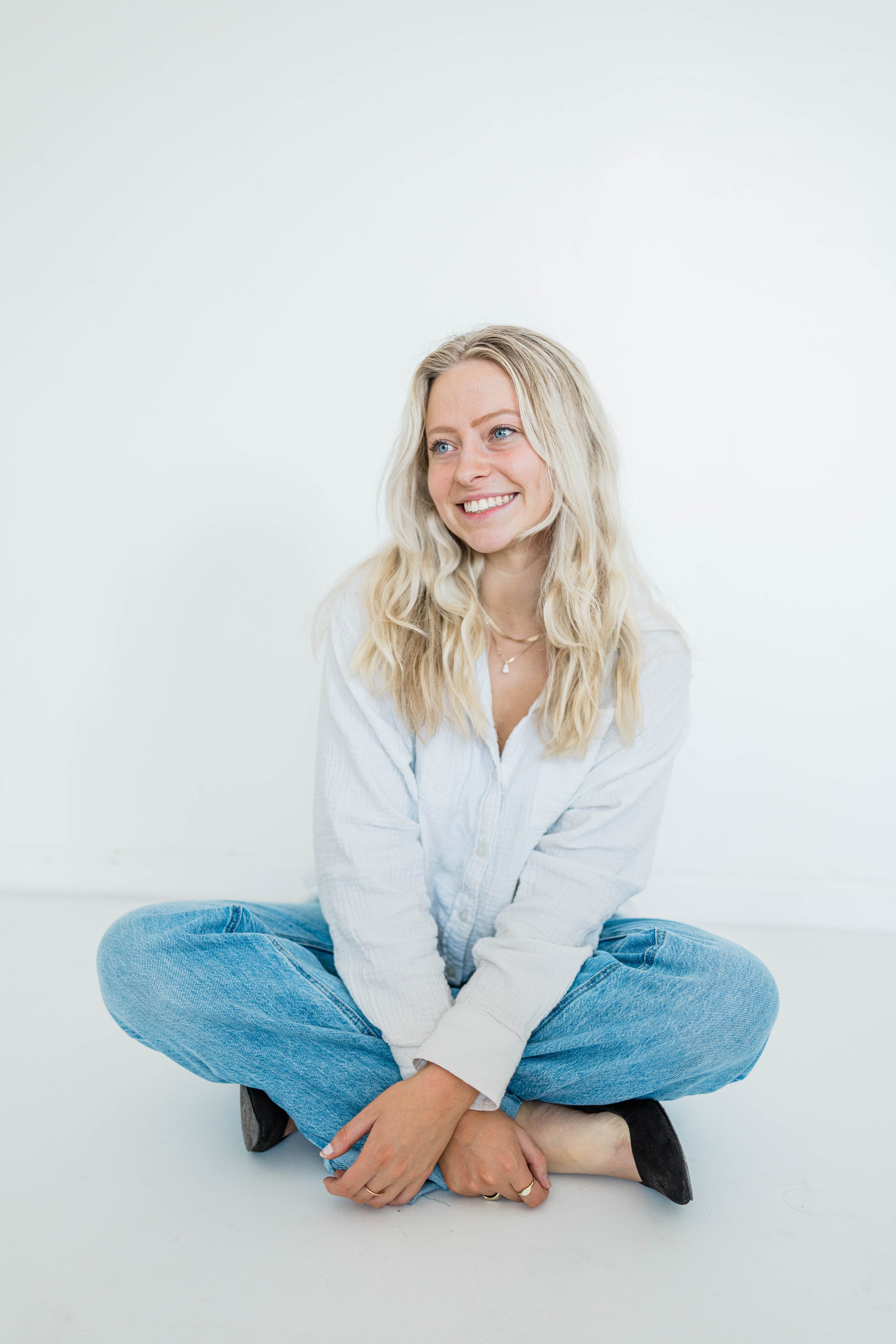 A woman sitting cross-legged on the floor against a plain white background, smiling and looking to her right. She is wearing a white loose shirt and blue jeans.
