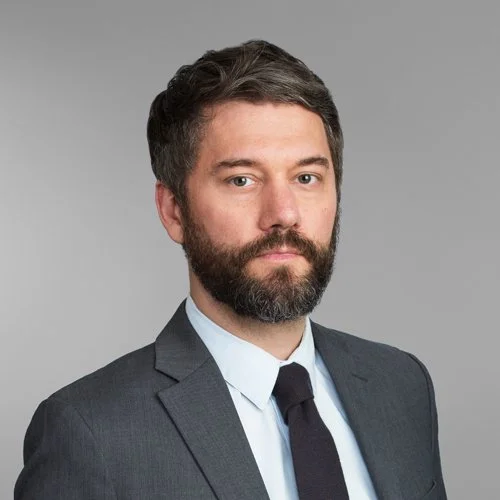 A man with dark hair and a beard dressed in a dark suit, white shirt, and black tie standing against a gray background.