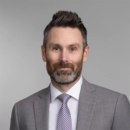 Professional portrait of a man in a gray suit with a white shirt and patterned tie, against a plain gray background.