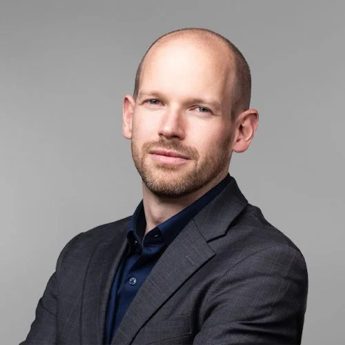 Professional portrait of a man with a bald head and beard, wearing a dark blazer and navy shirt, posing against a gray background.