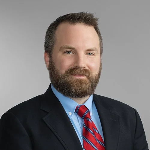 Professional man with beard, wearing a dark blazer, light blue shirt, and red striped tie, against a gray background.
