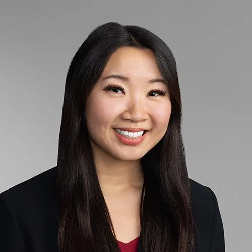Portrait of a smiling young Asian woman with long black hair wearing a black blazer against a gray background.