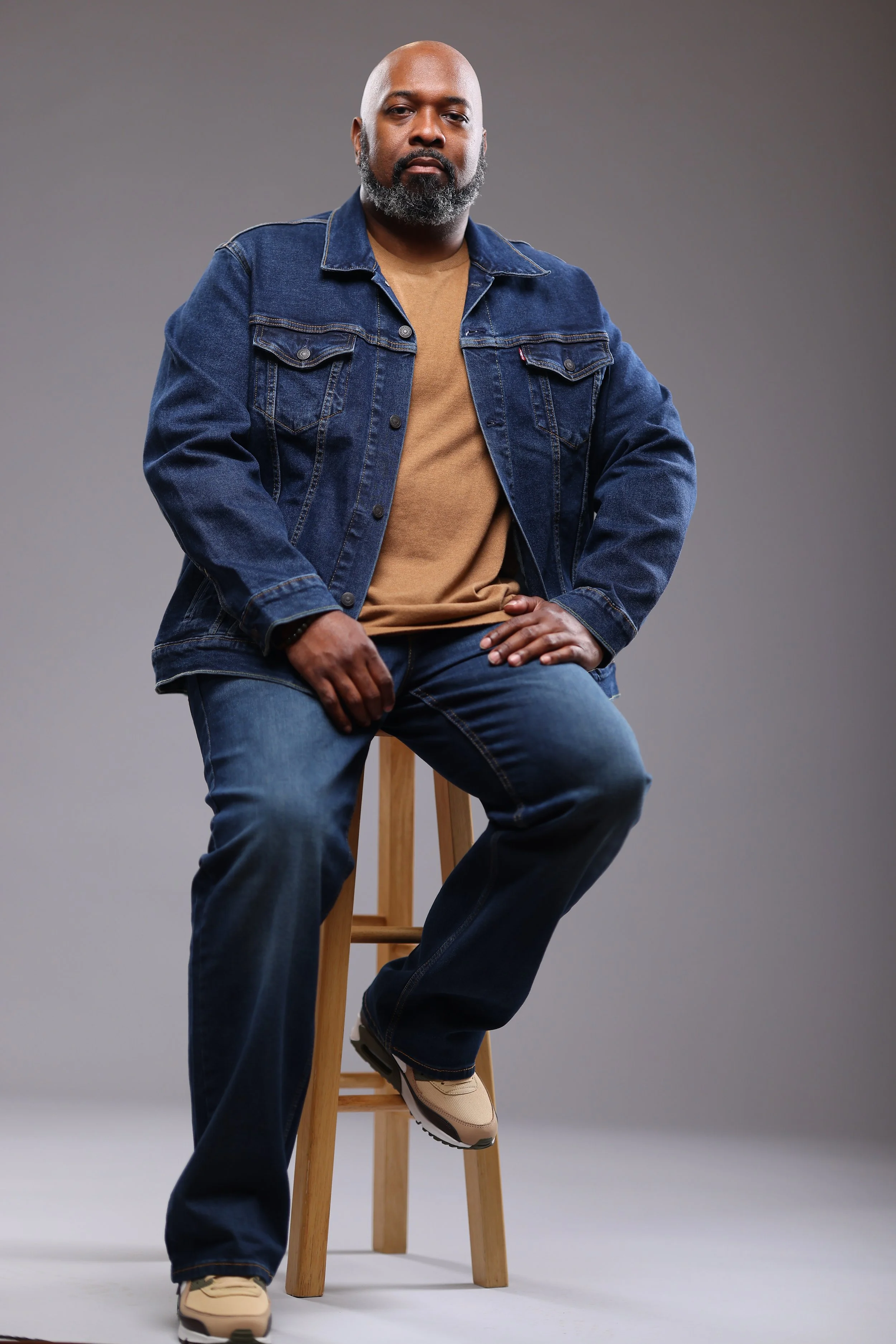 A man with a bald head and gray beard wearing a tan shirt, blue denim jacket, and jeans, sitting on a wooden stool against a plain gray background.
