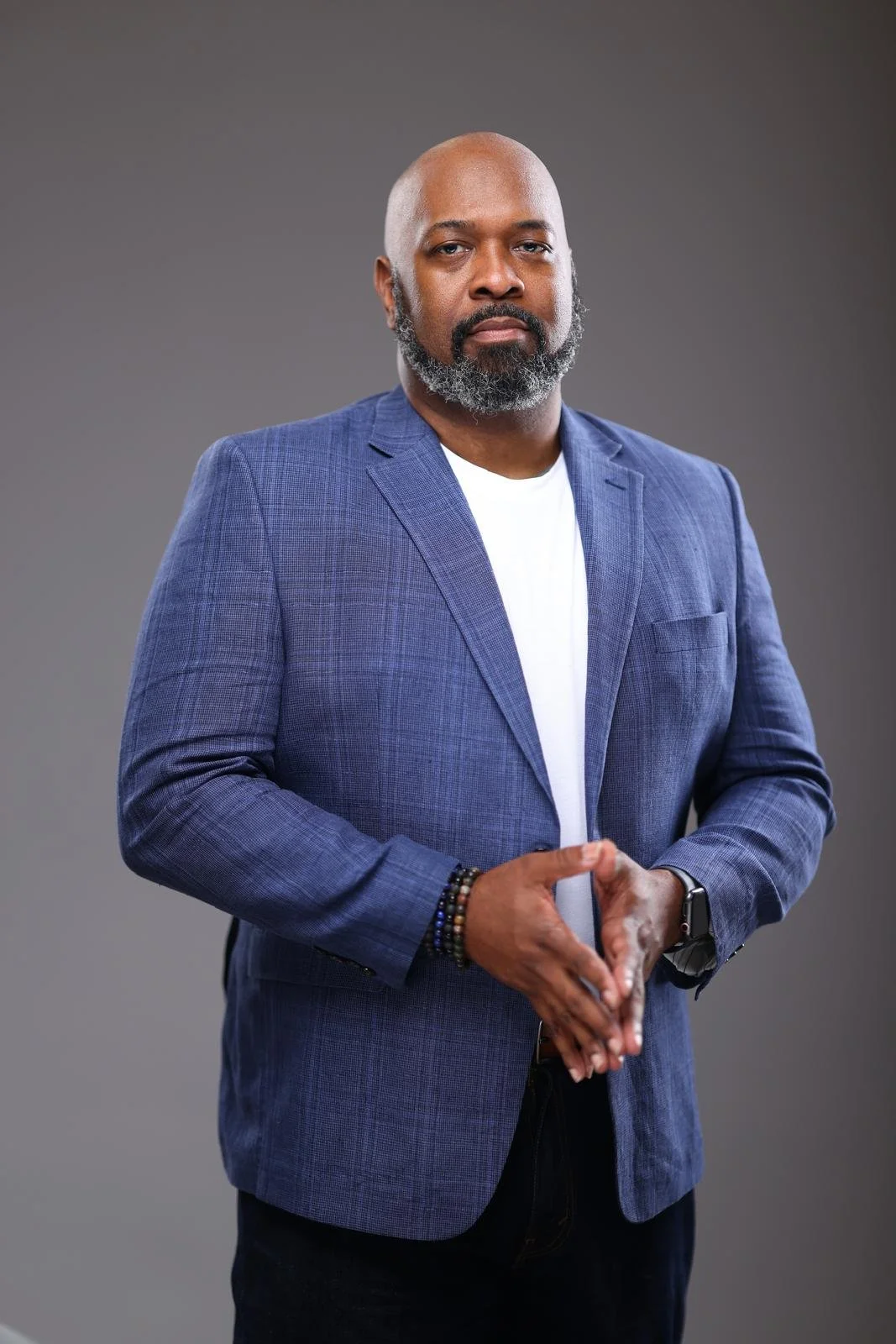 A man with a beard and mustache wearing a blue blazer over a white t-shirt, standing against a gray background.