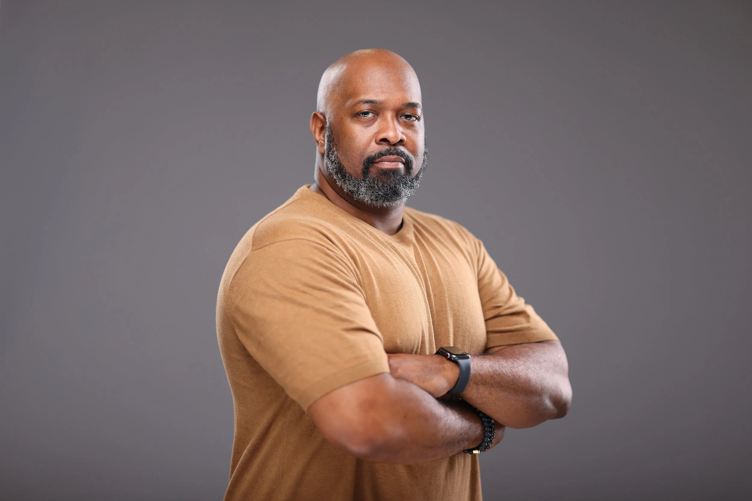 Portrait of a serious man with a beard and shaved head, wearing a tan T-shirt, crossing his arms, standing against a gray background.