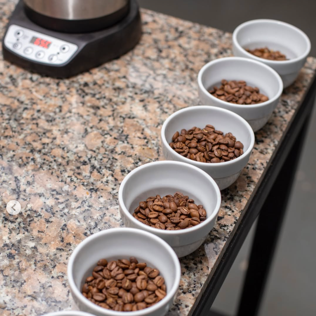 Five white bowls filled with roasted coffee beans placed on a granite kitchen countertop. Part of a coffee scale is visible in the background.
