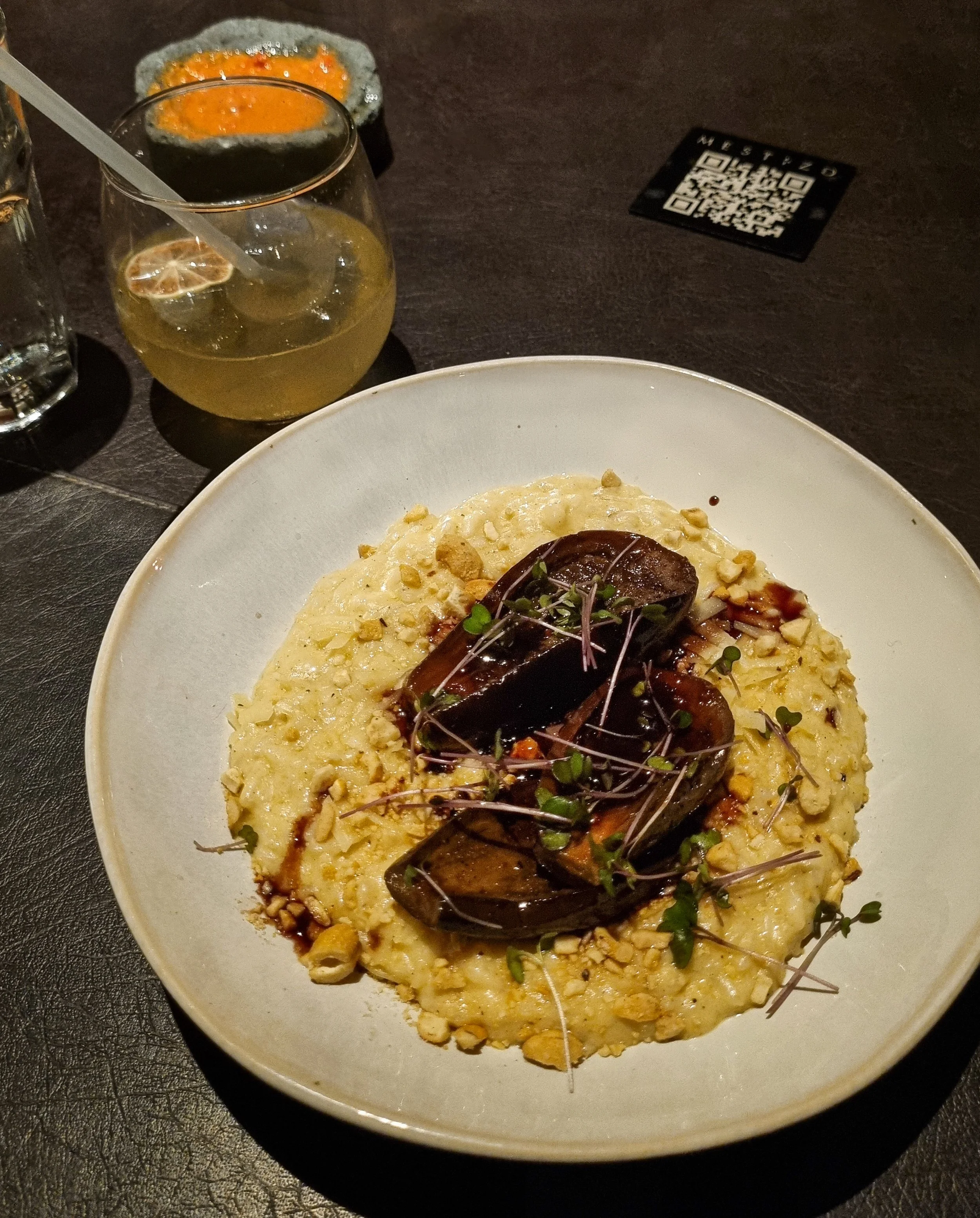 Close-up of a plated dish with creamy risotto topped with roasted vegetables and microgreens, with a dark background, drink, and QR code nearby.