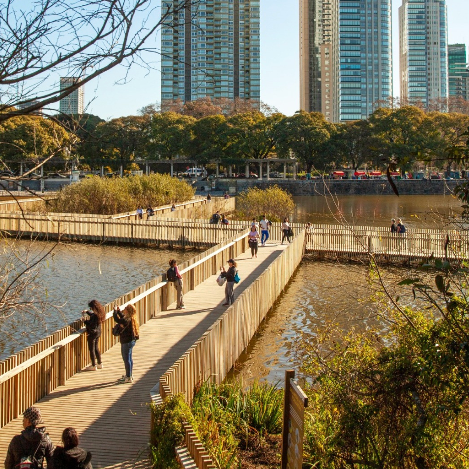 People walking along a wooden pier over water with tall modern buildings and trees in the background.
