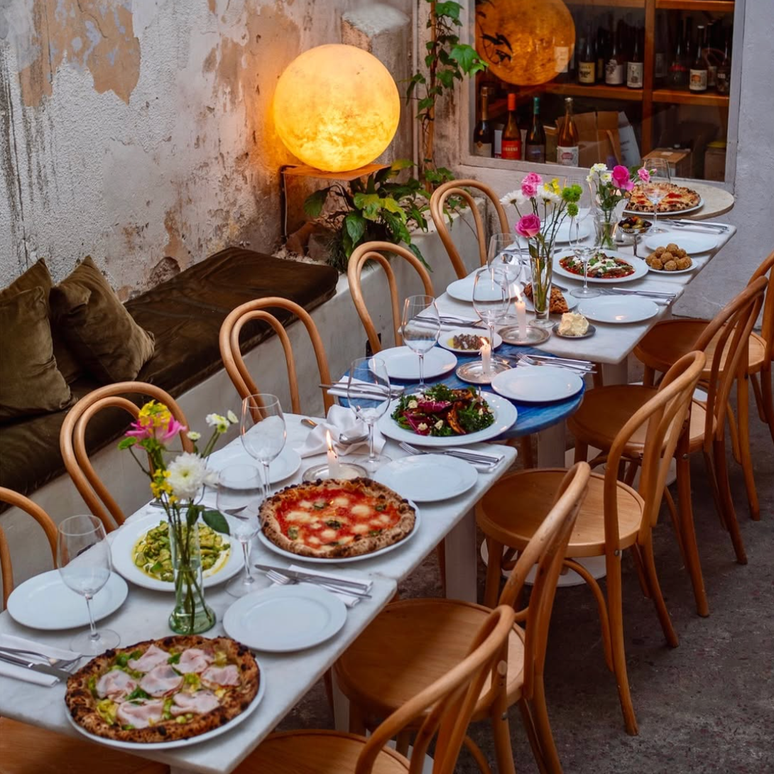 A dinner table set with pizzas, salads, flowers, wine glasses, and candles in a cozy restaurant with rustic walls and wooden chairs.