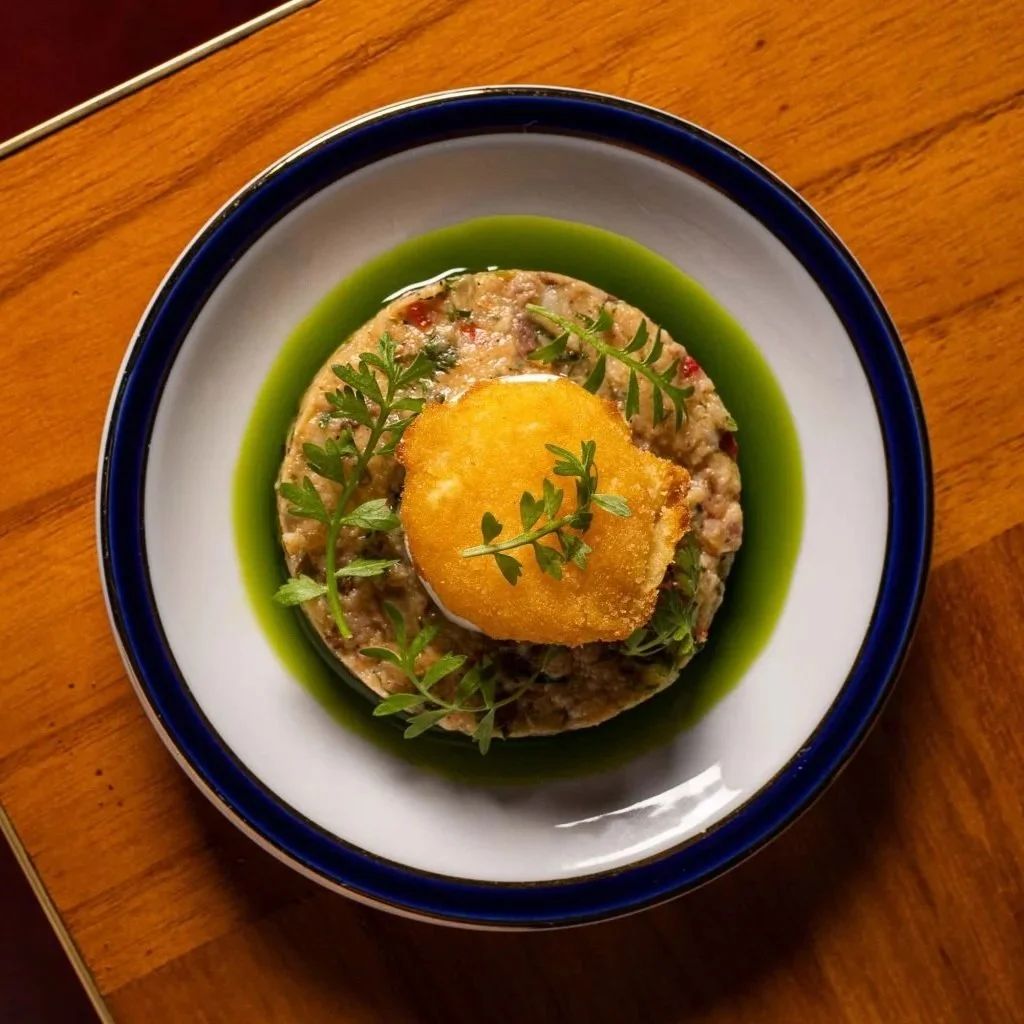A plated dish consisting of a mound of seasoned ground meat topped with a piece of fried plantain or sweet potato, garnished with fresh green herbs, and surrounded by green sauce or oil on a white plate with a dark rim, on a wooden table.
