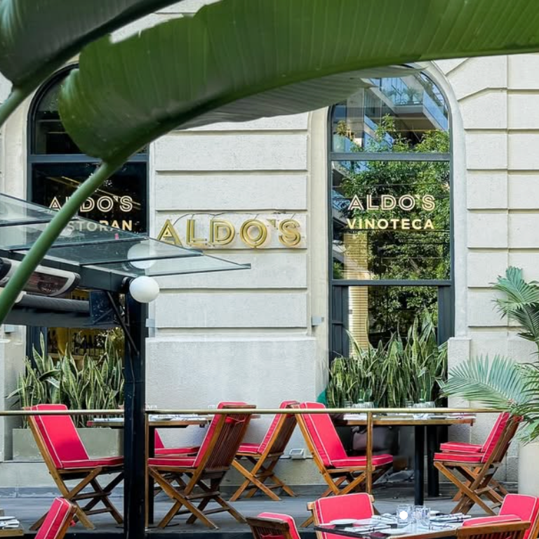 Outdoor seating area with wooden tables and red chairs at Aldo's Vinoteca restaurant, surrounded by green plants and a large building window reflecting trees.