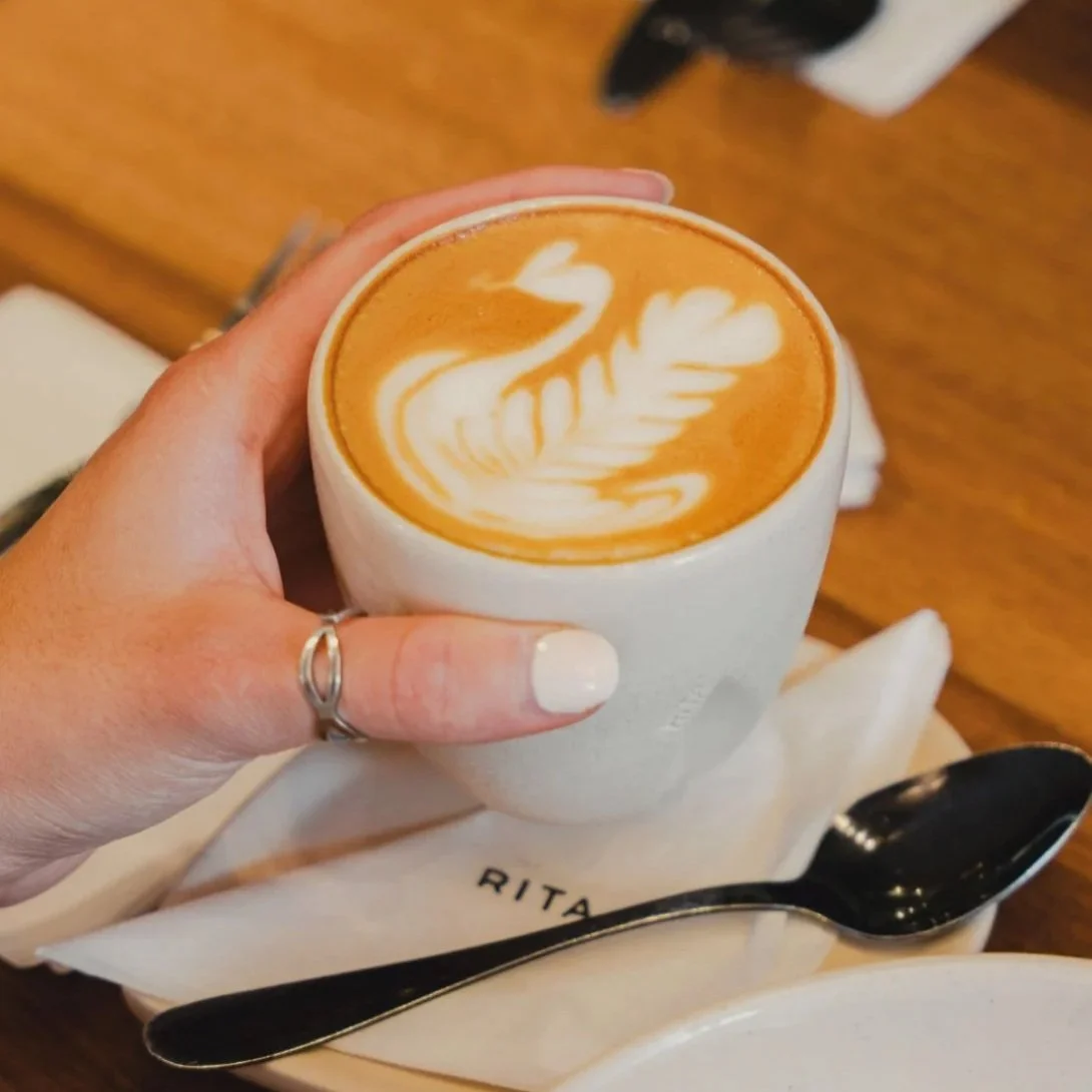 A hand holding a cup of coffee with latte art that resembles a swan or bird on top. The cup is white with a saucer, and there is a spoon on the saucer with a napkin underneath.