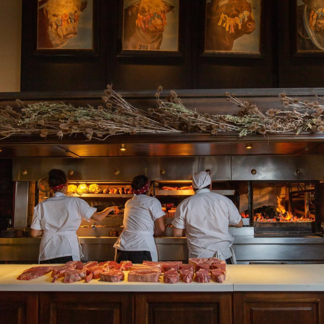 Three chefs in white uniforms and red bandanas working at a steakhouse grill with raw meat in front. Wood-fired grill with flames and cooked meats, dried herbs or flowers on the upper shelf, and framed animal portrait paintings on the wall.