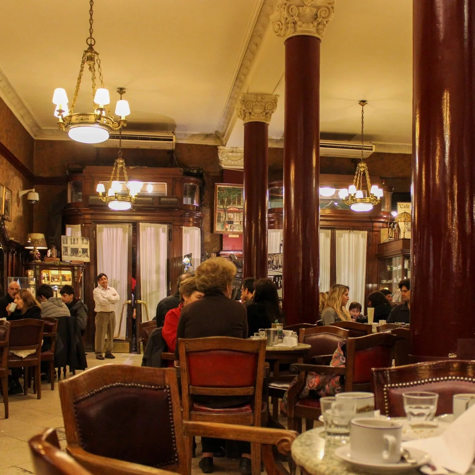 Interior of a vintage restaurant or café with old-fashioned wood furniture, large red columns, ornate chandelier lighting, and patrons seated at tables enjoying their meals.