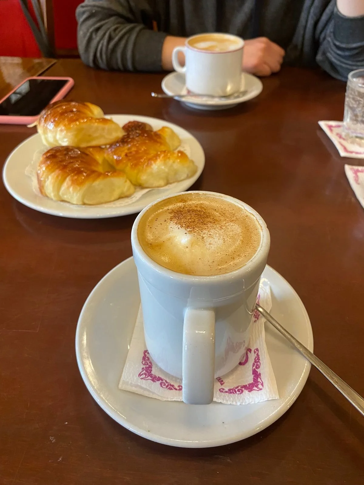 Two cups of coffee with foam, a plate of cinnamon rolls, and a person's arm at a wooden table in a cozy cafe setting.