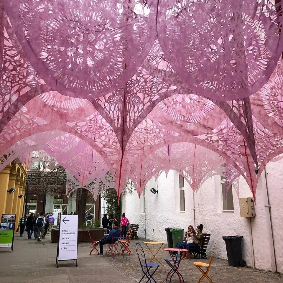People sitting on benches and walking under colorful pink and purple paper lanterns hanging overhead in an outdoor plaza.